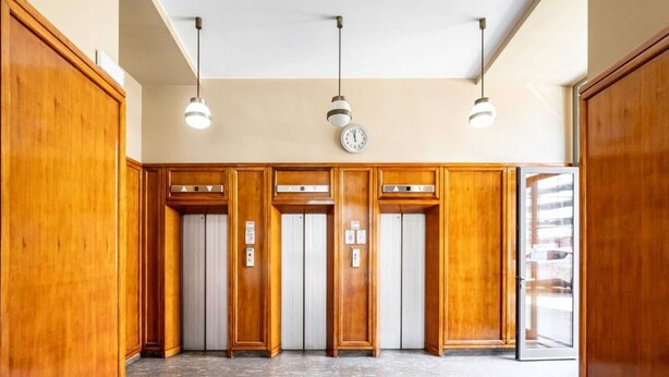 Una hall con pannelli in legno, tre lampadari a sospensione, un orologio e una porta vetrata che dà sull'esterno.