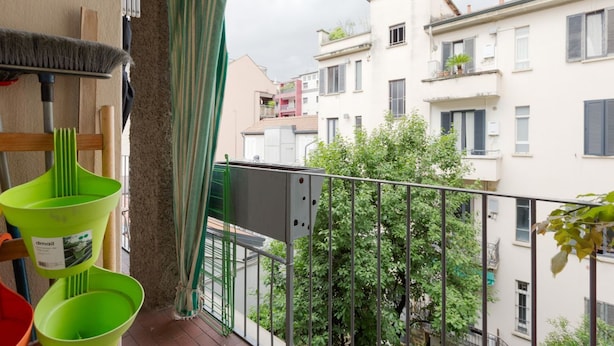 A balcony with a view of neighboring buildings, a metal railing, and a green plant. - GuestHost - APT in the NOLO neighborhood! 500m from the M1 subway! (Milano)