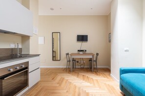 Une cuisine moderne dotée d’un four encastré, une table à manger avec des chaises, un téléviseur à écran plat fixé au mur et un canapé bleu dans l’espace salon.