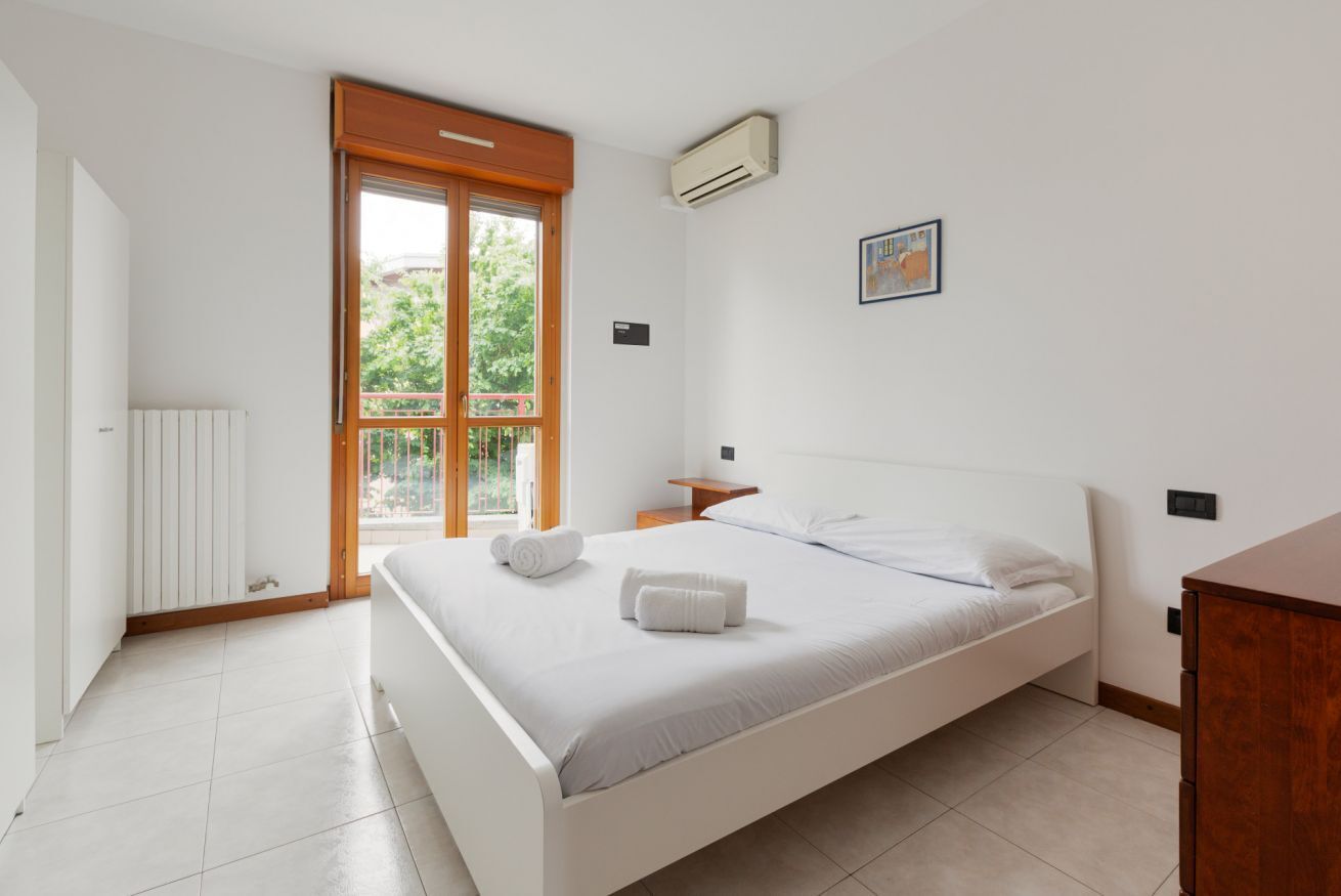 A bedroom with a bed, a wooden door, a window, an air conditioner, a bedside table, and a wall-mounted light switch.