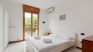 Ein Schlafzimmer mit einem Bett, einer Holztür, einem Fenster, einer Klimaanlage, einem Nachttisch und einem an der Wand montierten Lichtschalter.