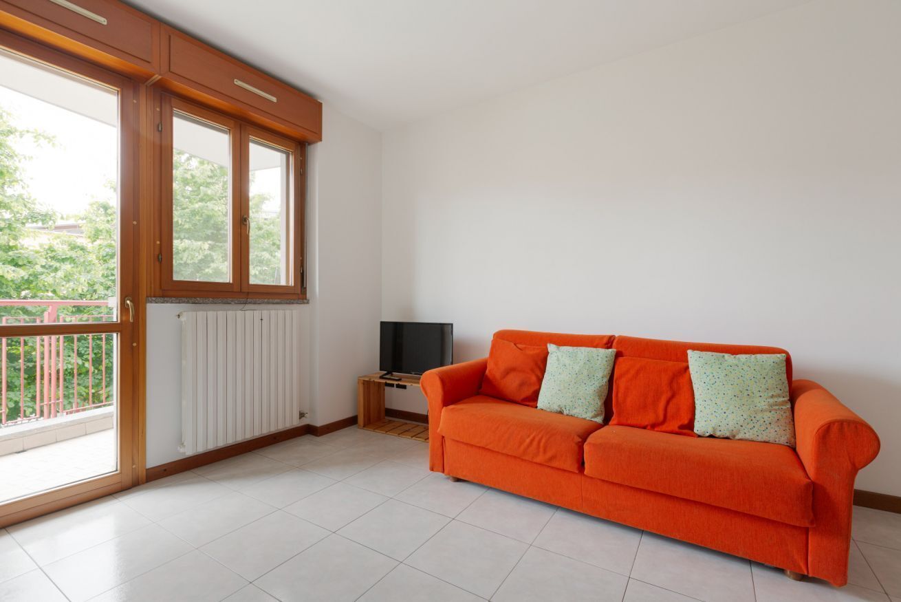 Ein Zimmer mit großem Fenster, einem weißen Heizkörper, einem Fernseher auf einem Holzständer und einem orangefarbenen Sofa mit gemusterten Kissen.