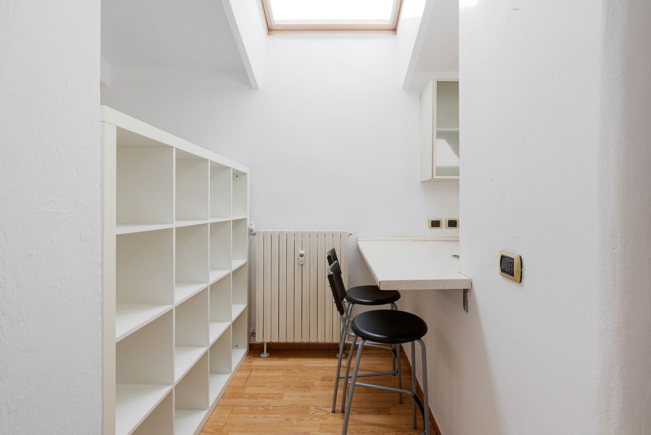 A narrow room with white walls, a wooden floor, a skylight, a radiator, a white shelf unit, and two black chairs.