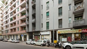 Ein mehrstöckiges Gebäude mit Balkonen, eine Straße mit parkenden Autos und Geschäften, darunter eine Pizzeria.