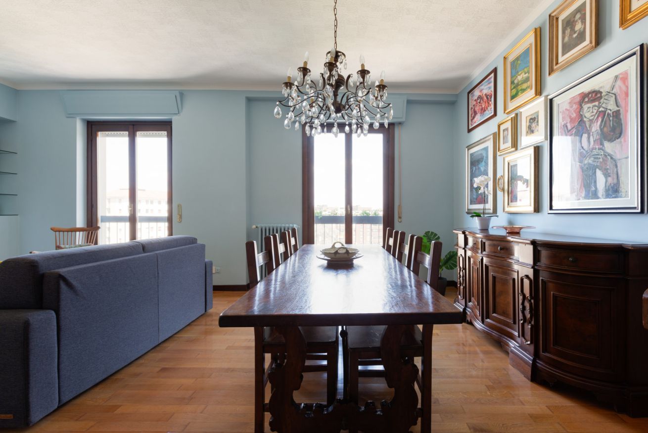 Uma sala de jantar com uma mesa de madeira, cadeiras, um candelabro, um sofá azul e um armário com quadros.