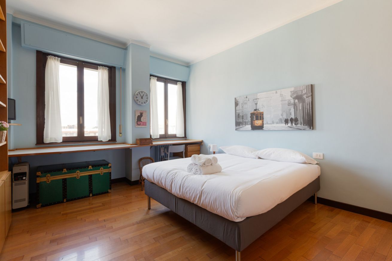Quarto com cama, escrivaninha, cadeira, janela com cortinas, relógio de parede, quadro emoldurado e um baú verde.