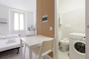 A compact hotel room with a bed, a small table, a chair, a washing machine, a toilet, and a wooden panel wall.