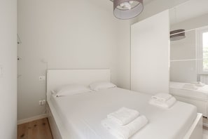 A clean, white room with a bed, a mirror, and a window.