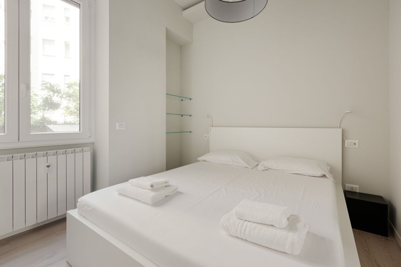 A bedroom with a bed, a window, a radiator, and a wall-mounted shelf.
