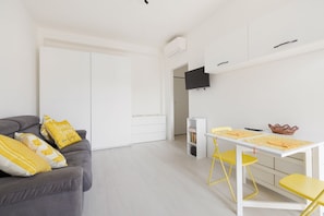 A modern living room with a grey sofa, yellow cushions, a white dining table with yellow chairs, a flat-screen TV mounted on the wall, and a white wardrobe.