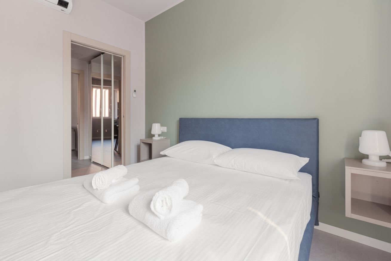 A neatly made bed with white linens and towels, a blue headboard, a bedside table with a lamp, and a mirror on the wall.