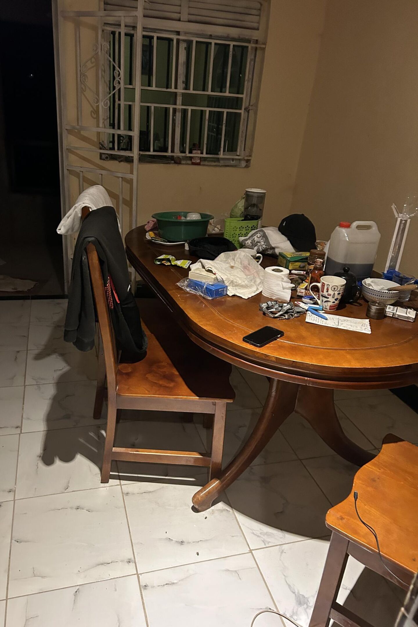 Een rommelige eettafel met verschillende voorwerpen, een stoel en een raam met tralies.