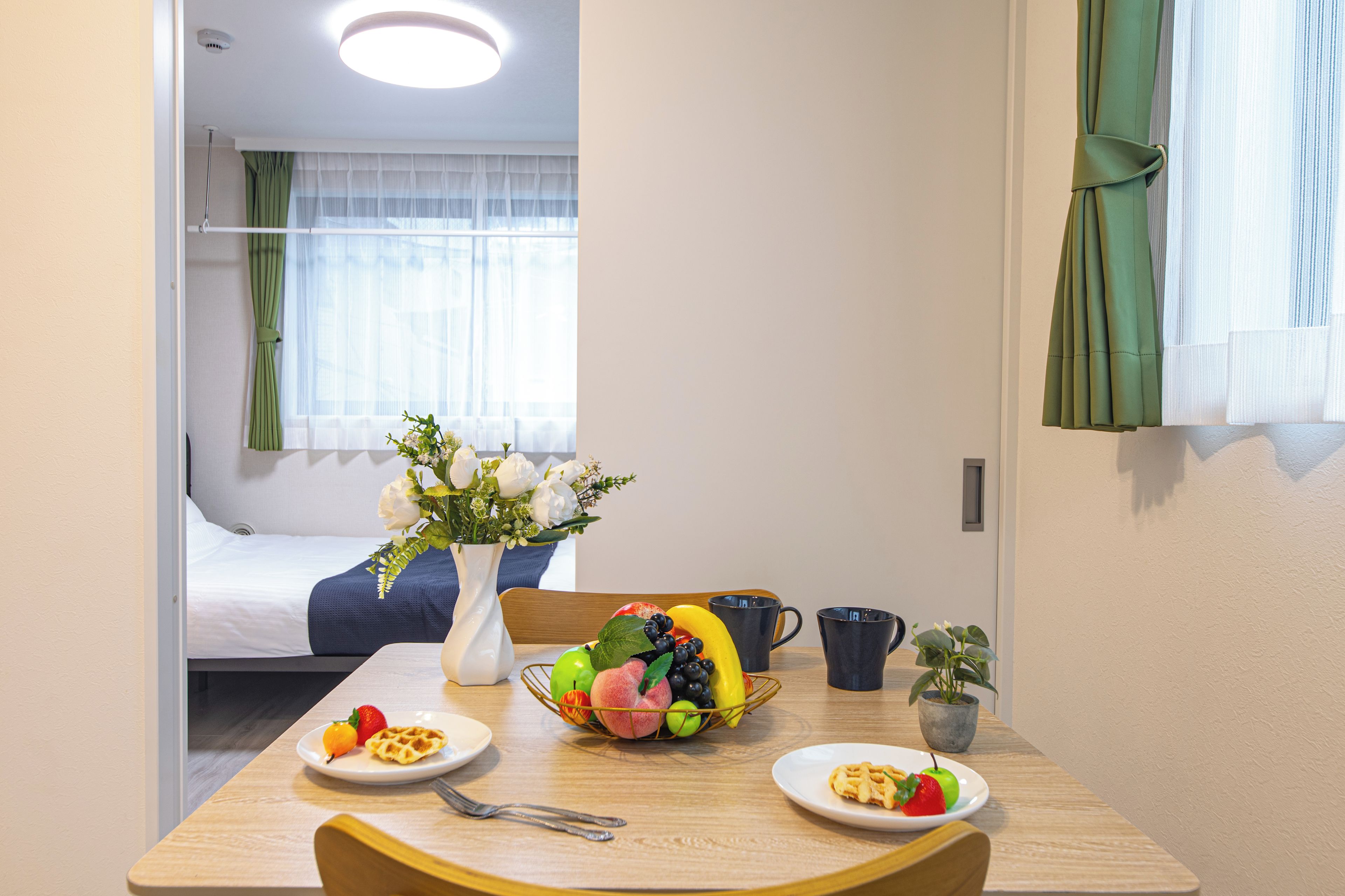 Hotellihuone, jossa on sänky, aamiaistarvikkeilla katettu ruokapöytä, maljakko kukkineen ja näkymä huoneeseen ikkunasta.