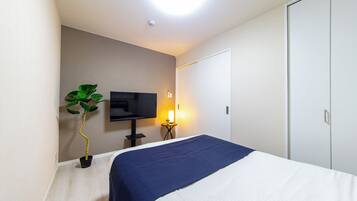 A hotel room with a bed, a television, a bedside table with a lamp, and a potted plant.