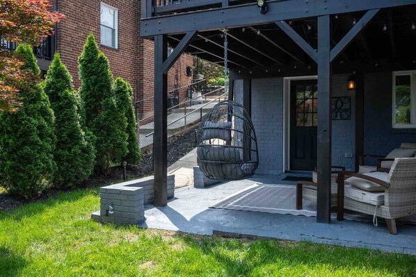 A covered outdoor seating area with a hanging chair, a brick wall, and a staircase leading to an upper level.