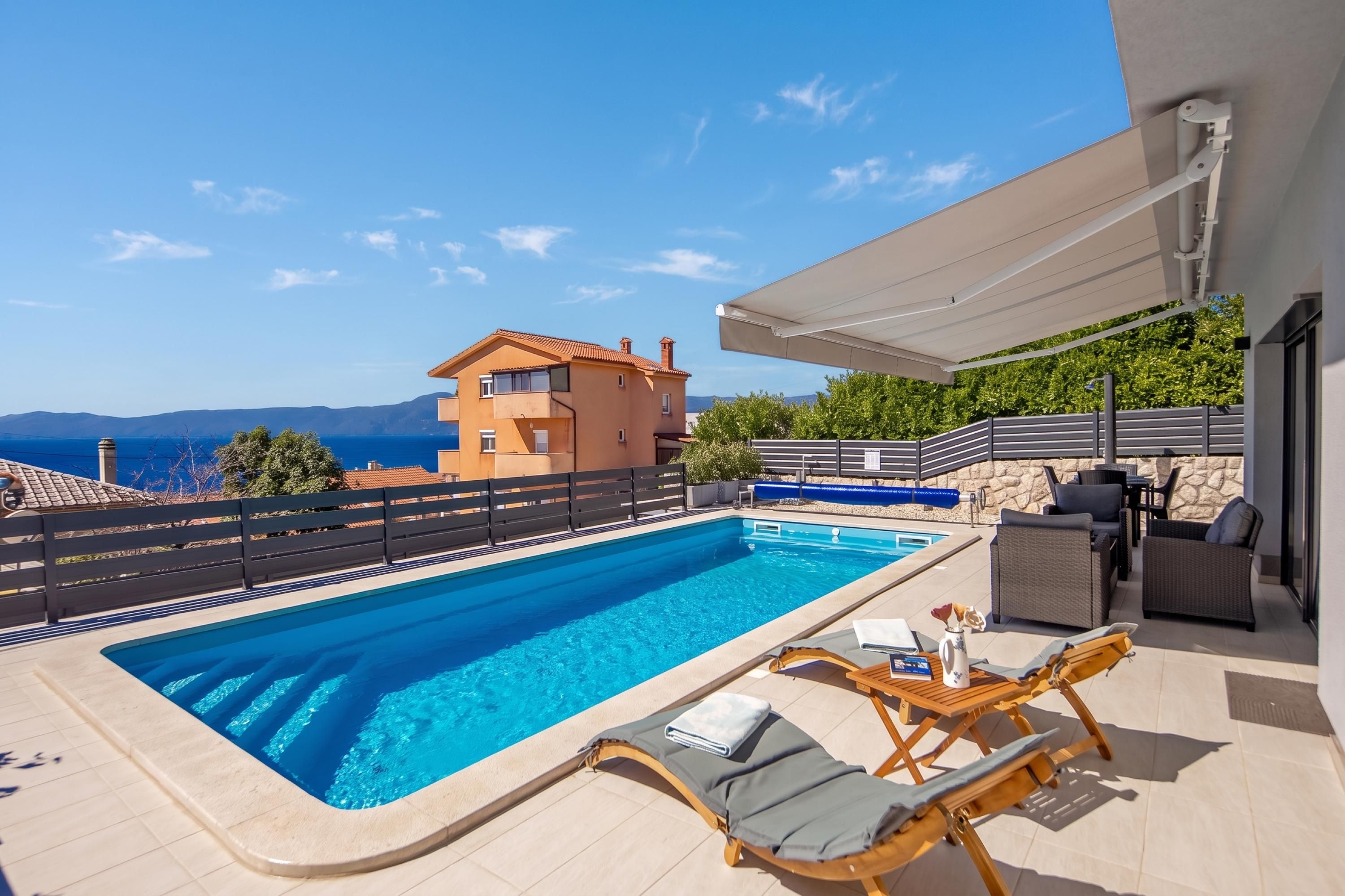 En swimmingpool med liggestole og et bord, en overdækket terrasse samt hav- og bjergudsigt.