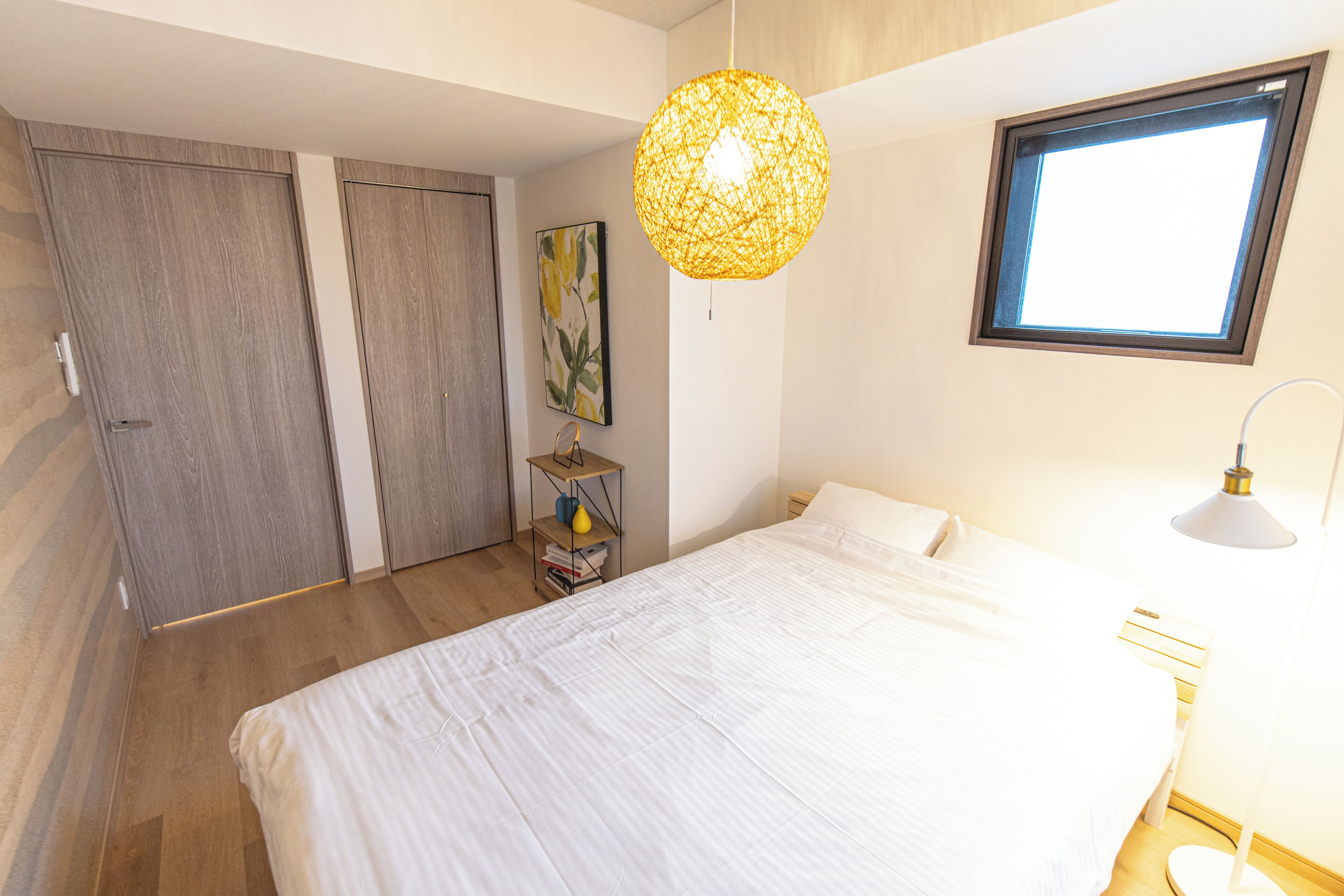A bedroom with a bed, a bedside lamp, a window, a wall-mounted shelf with books, and a door.