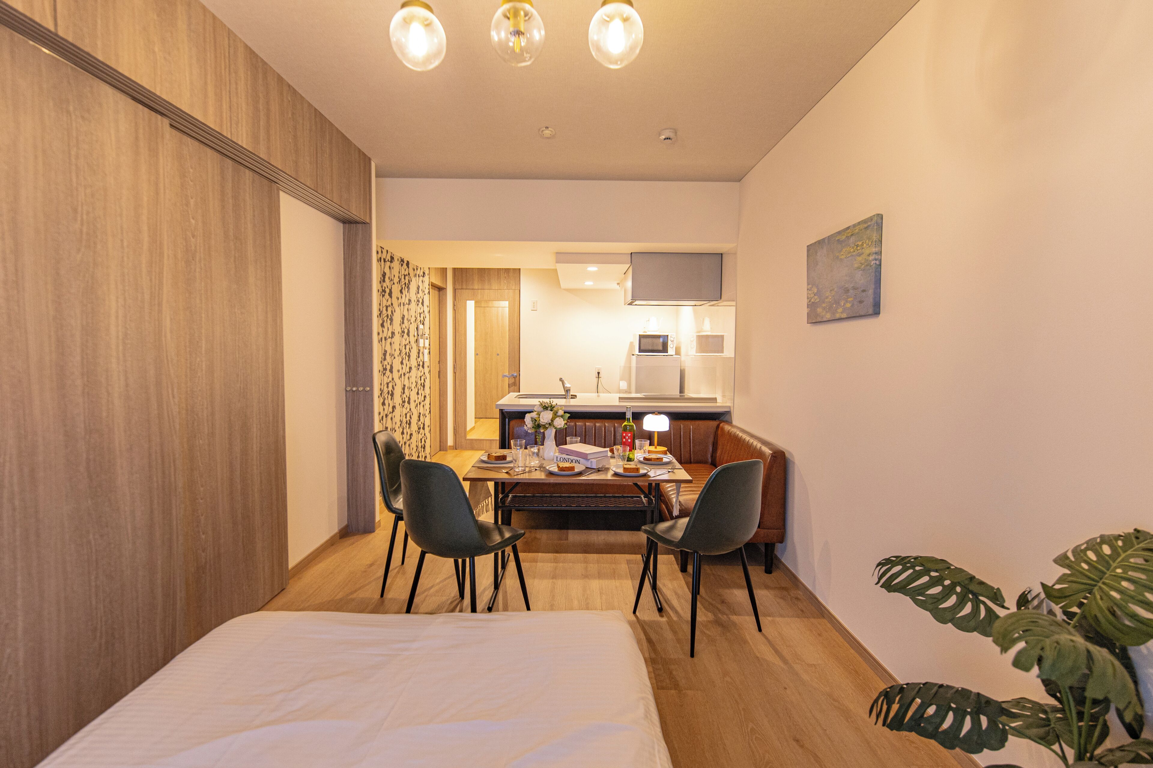 Habitació d'hotel moderna amb un llit, una taula de menjador amb cadires, una cuineta i una planta gran.