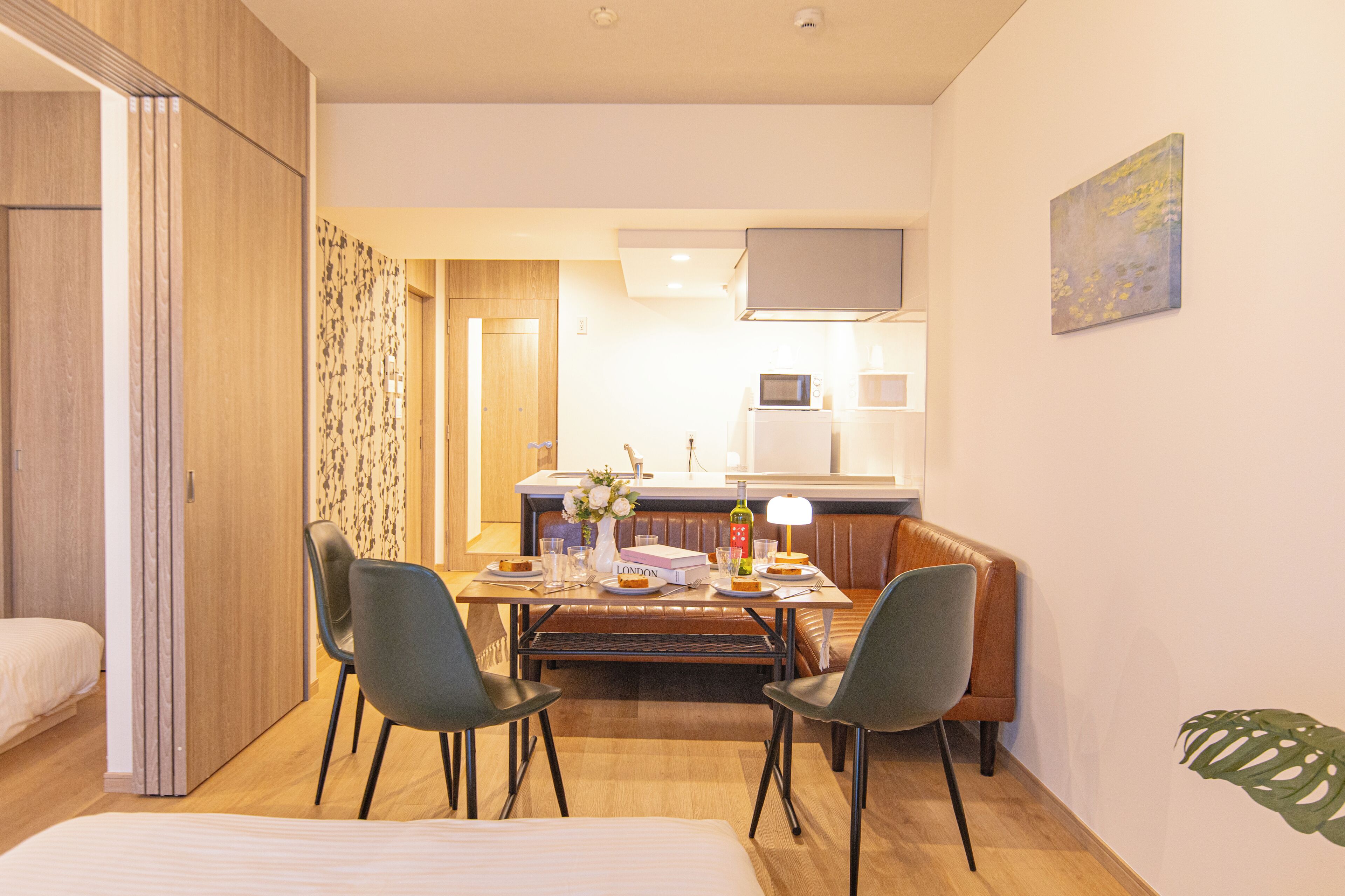 A modern hotel room with a wooden wardrobe, a dining table with chairs, a small kitchen area, and a single bed.