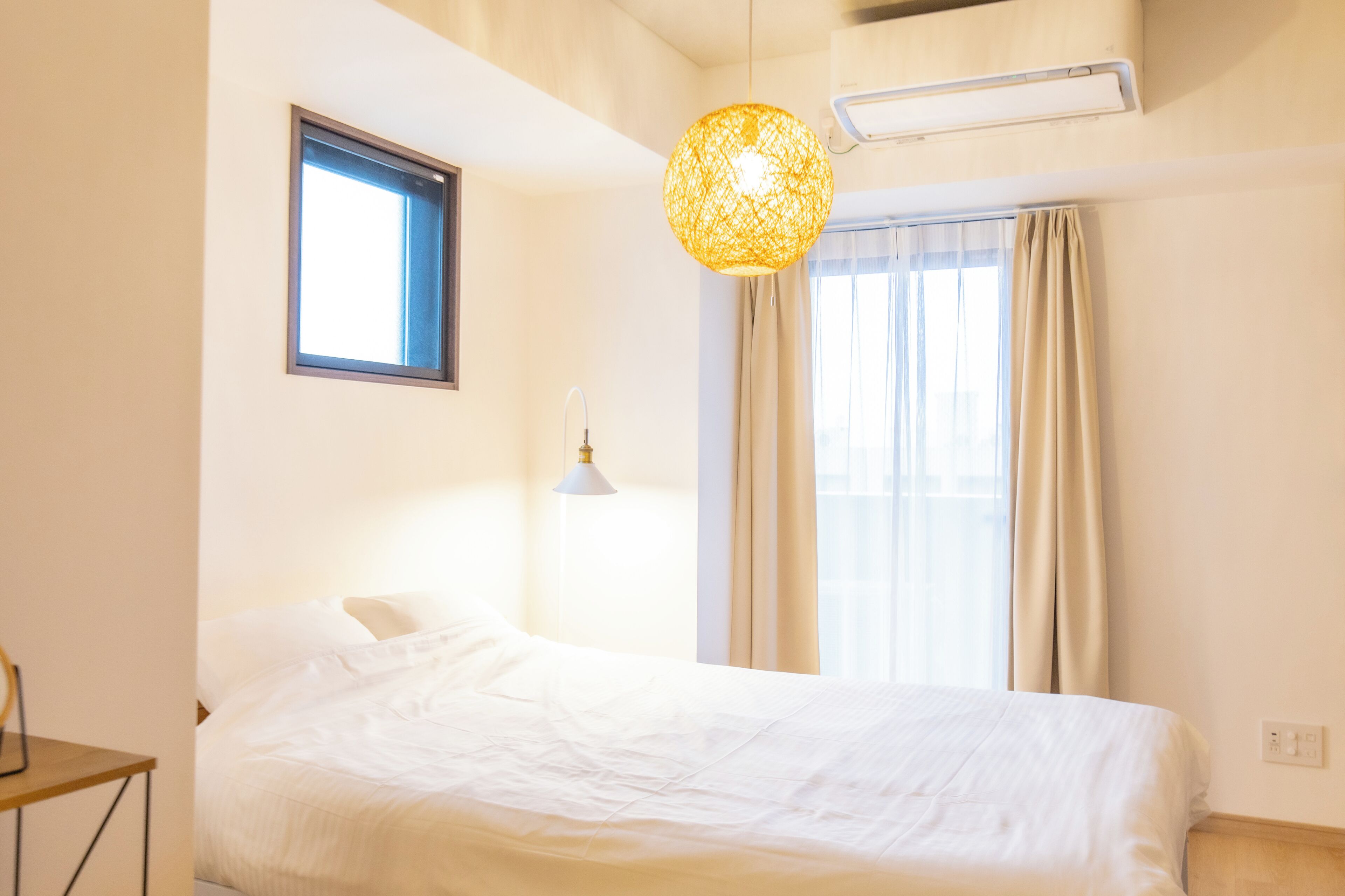 Habitación de hotel con cama, ventana con cortinas, aire acondicionado de pared y lámpara colgante.