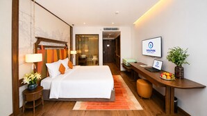 A hotel room with a bed, bedside table, lamp, desk, laptop, and a television. - Apec Mandala Mui Ne by Dolphin Hospitality (Mui Ne)