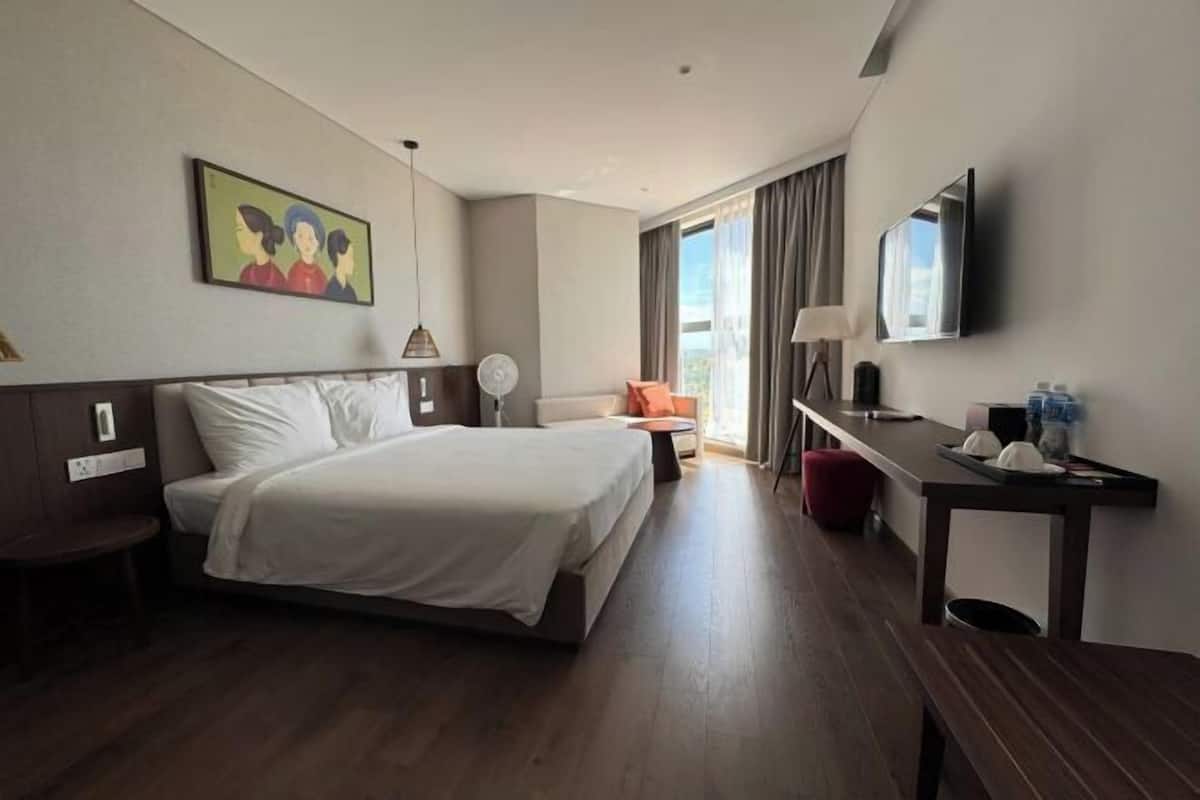 A hotel room with a bed, a sofa, a desk, a TV, and a painting on the wall.