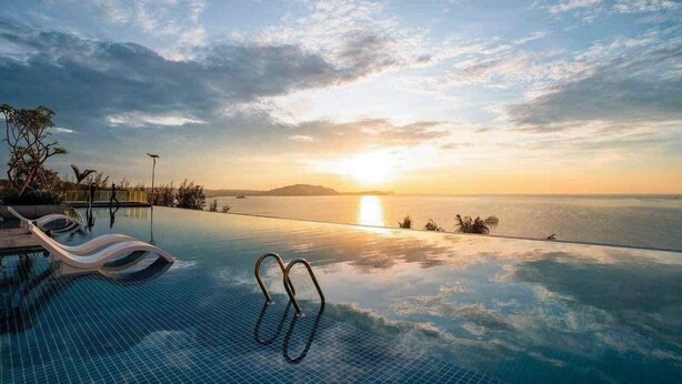 An infinity pool with lounge chairs, a clear sky, and a sunset over the ocean. - Apec Mandala Mui Ne by Dolphin Hospitality (Mui Ne)