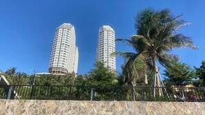 Two tall, modern buildings with a stone wall in the foreground and palm trees. - Apec Mandala Mui Ne by Dolphin Hospitality (Mui Ne)