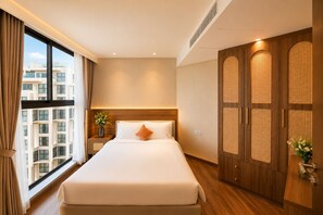 A hotel room with a large bed, a wardrobe with a woven design, a nightstand with a vase, and a view of a building through the window. - Apec Mandala Mui Ne by Dolphin Hospitality (Mui Ne)