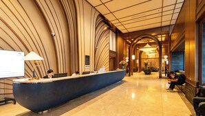 A reception area with a curved reception desk, a flat-screen TV, and a seating area with a person sitting on a bench. - Apec Mandala Mui Ne by Dolphin Hospitality (Mui Ne)