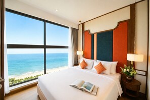 A hotel room with a large window overlooking a beach, a bed with white linens, two bedside lamps, and a small table with a vase of flowers. - Apec Mandala Mui Ne by Dolphin Hospitality (Mui Ne)