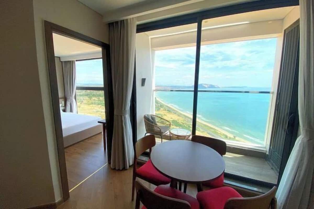 A room with a bed, a dining table with chairs, and a large window offering a view of the sea.
