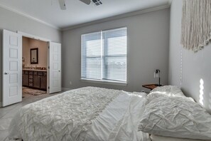 Ein Schlafzimmer mit einem großen Bett, einem Fenster mit Jalousien, einem Nachttisch mit Lampe und einer offenen Tür zu einem Badezimmer.