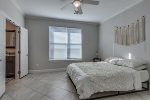 Un dormitorio con una cama, un ventilador de techo, una ventana con persianas y una decoración de macramé en la pared.