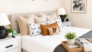A neatly arranged bedroom with a bed, pillows, a bedside table with a lamp, a wall-mounted hat rack, and a framed picture.