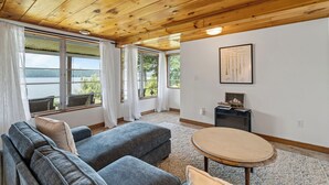 Une pièce chaleureuse avec un plafond en bois, de grandes fenêtres avec des rideaux, un canapé gris, une table ronde et un tableau encadré au mur.