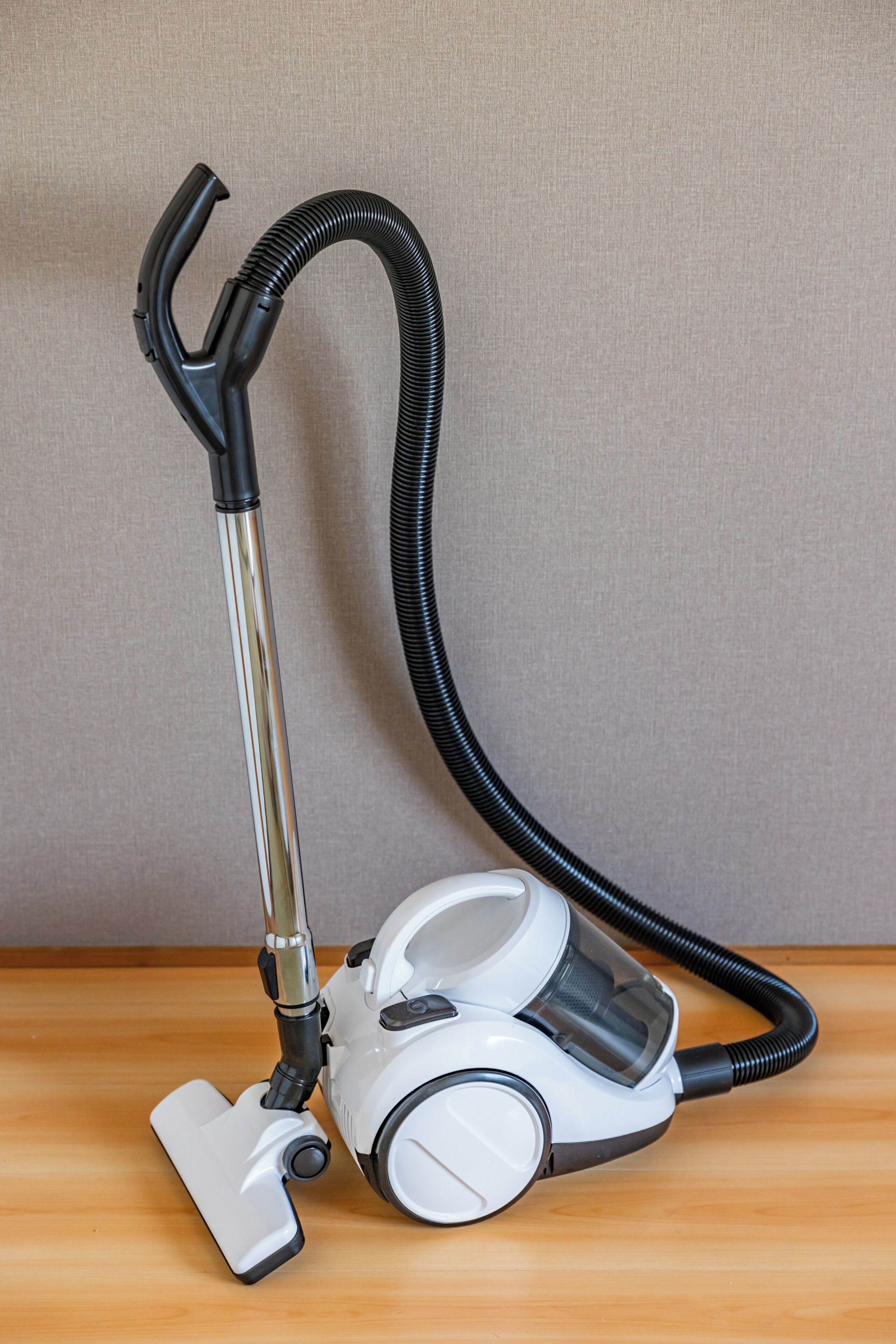 A vacuum cleaner with a long hose and a compact body, placed on a wooden surface against a plain wall.