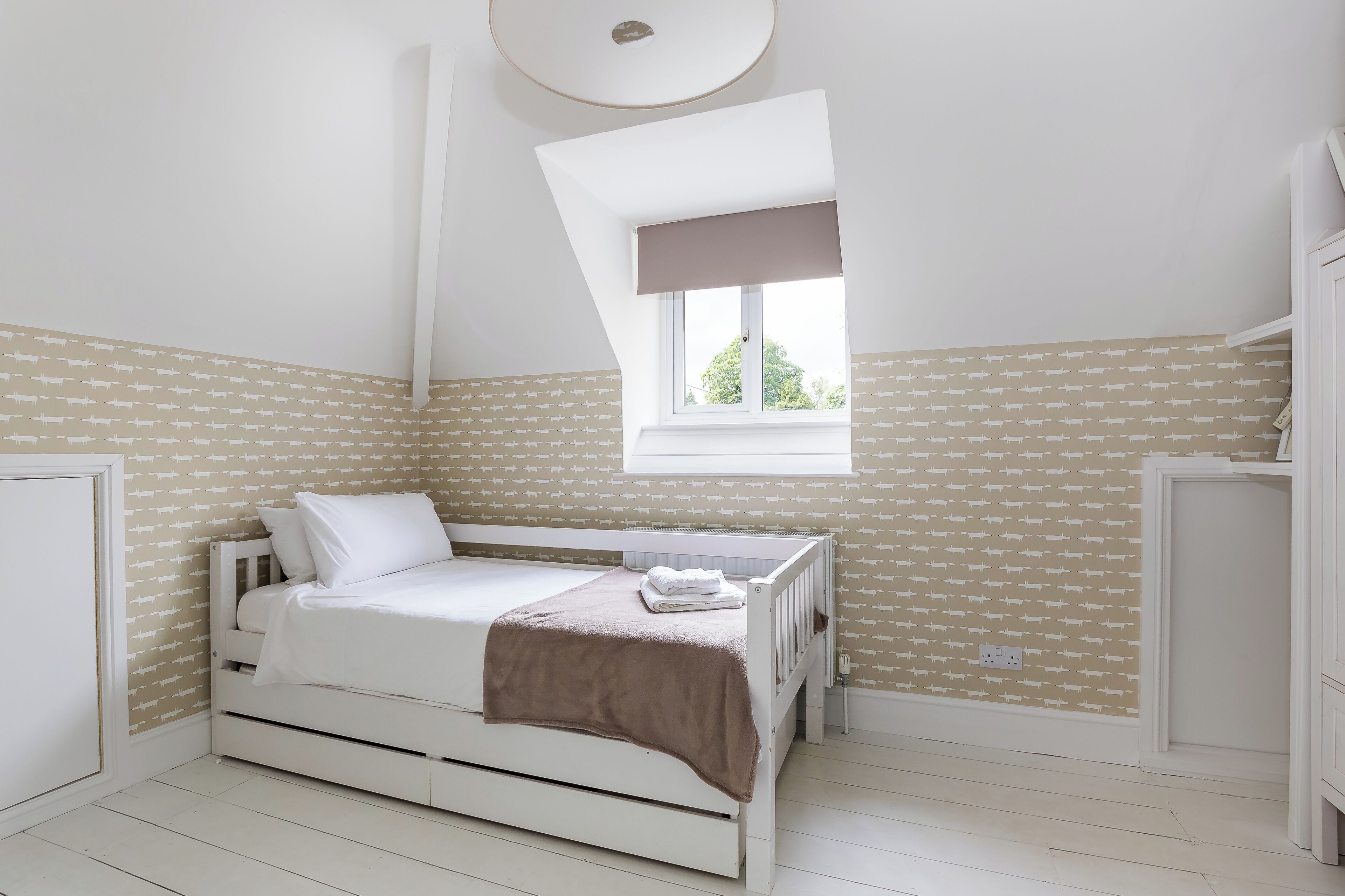 Un dormitorio con techo inclinado, una ventana con persiana marrón, una cama con ropa blanca y papel tapiz estampado.