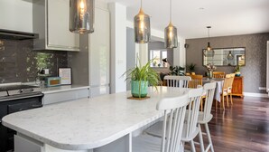 Una cocina moderna con encimera de mármol, iluminación colgante y una zona de comedor con sillas blancas y una mesa de madera.