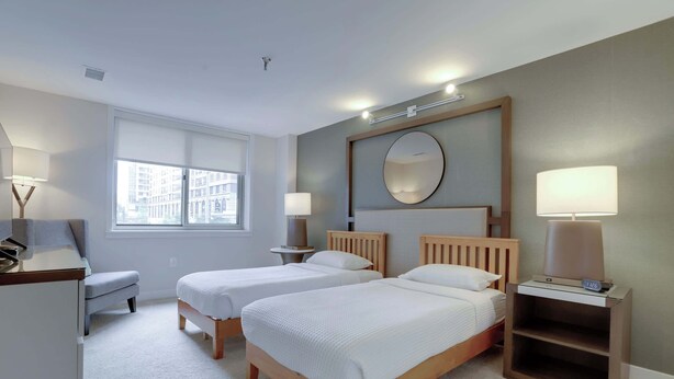 A hotel room with two single beds, a desk, a chair, a bedside table with a lamp, and a large window offering a city view.