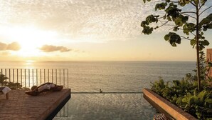 A wooden deck with a glass-bottomed infinity pool overlooking the sea, a wicker basket, and a person swimming. - Rosa Morada: Luxury Villas with Private Beach (San Francisco)