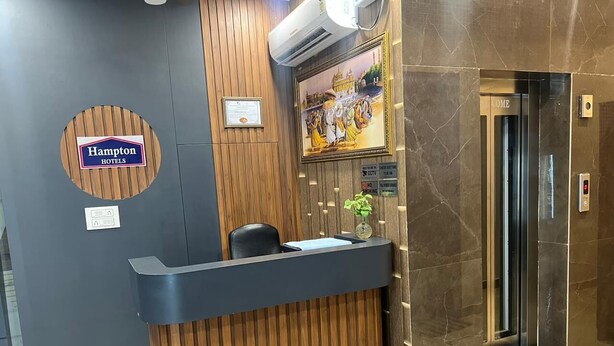 A hotel reception desk with a wooden panel design, a Hampton Hotels sign, a framed picture of a historical scene, and an elevator. - Om B & B (Amritsar)
