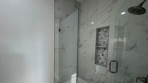 Un baño moderno con ducha acristalada, baldosas de mármol y un separador de vidrio con diseño de rombos.