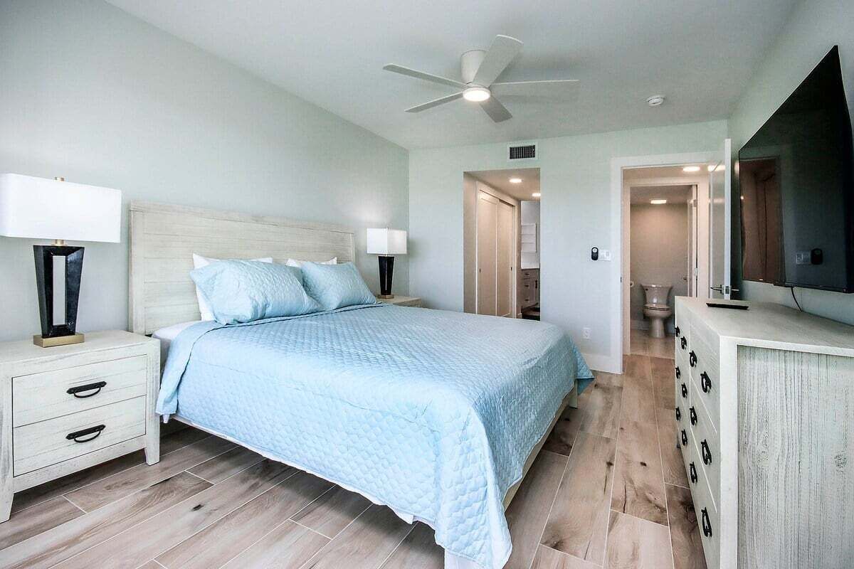 Un dormitorio con cama, mesitas de noche, ventilador de techo, cómoda y televisor.