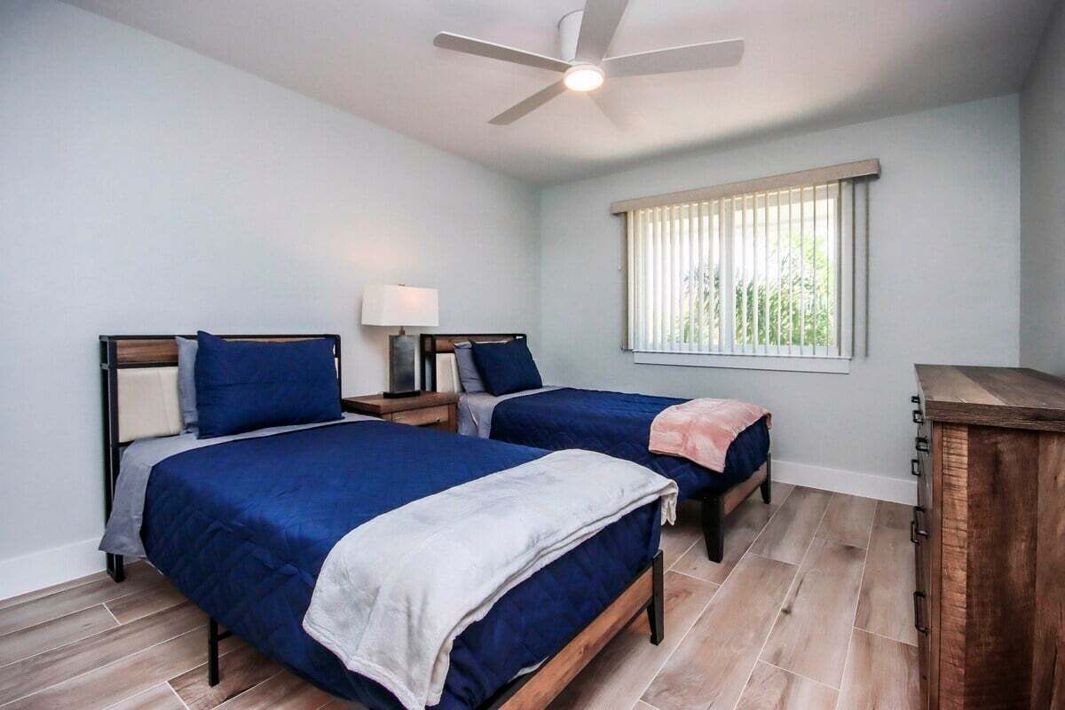 Une chambre avec deux lits, un ventilateur de plafond, une fenêtre avec des stores et une armoire en bois.