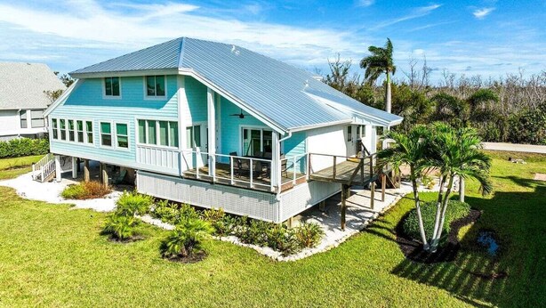 A two-story house with a blue roof and white exterior, featuring a covered porch and a small garden area with palm trees.