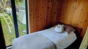 Ett litet, ombonat sovrum med en sänggavel i trä, en enkelsäng med ett vitt täcke och ett fönster med utsikt över en trädgård.