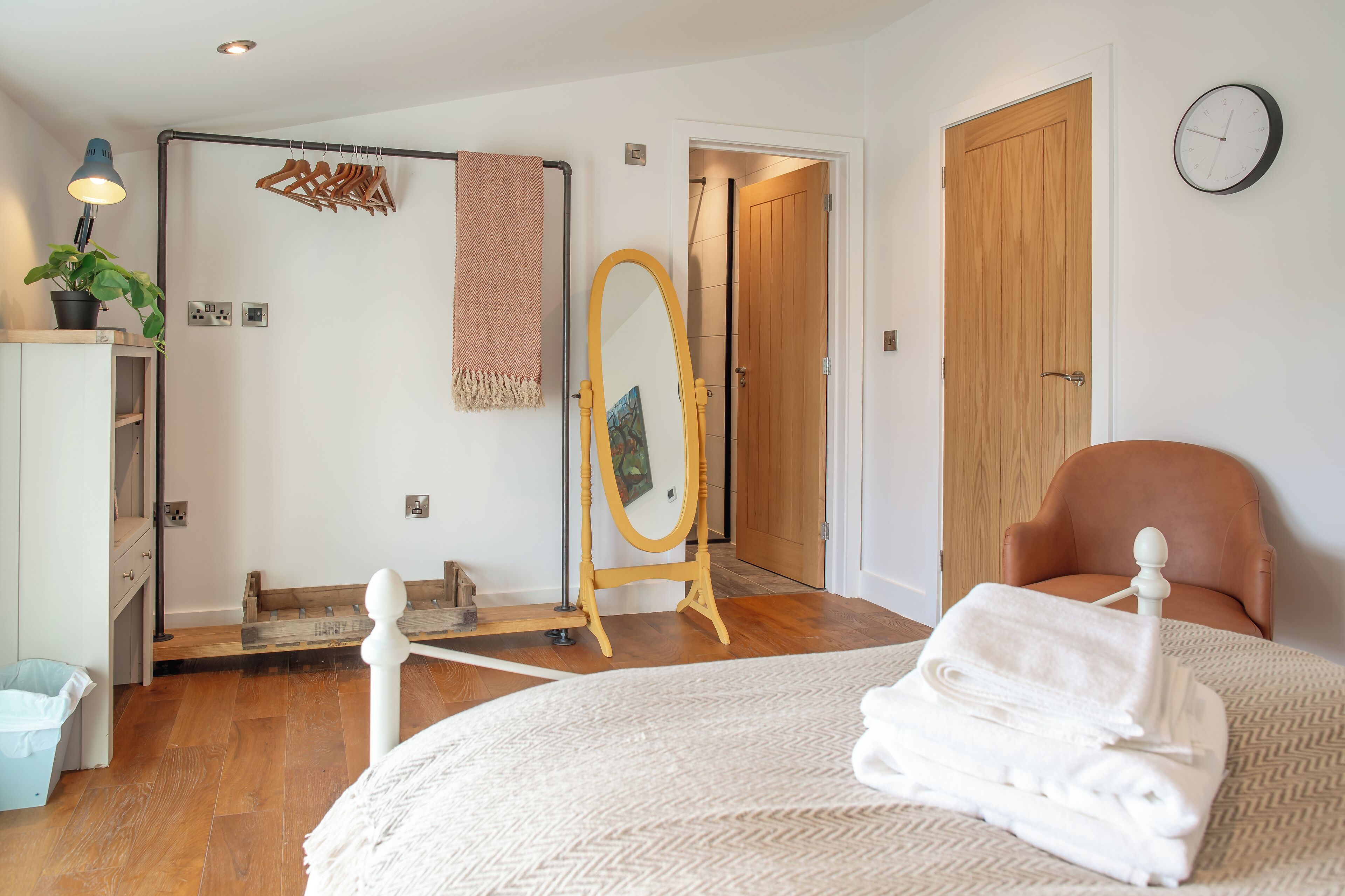 A bedroom with a bed, a wooden floor, a mirror, a chair, and a door.