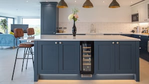 A modern kitchen with dark blue cabinets, a central island, pendant lighting, and a wine cooler. - Orchard Barn-Luxury 4 BD Barn Conversion (Pershore)