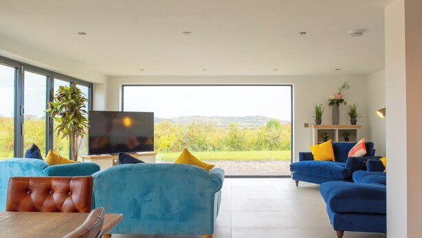 A spacious living room with large windows, a television, and two sofas with colorful cushions. - Orchard Barn-Luxury 4 BD Barn Conversion (Pershore)
